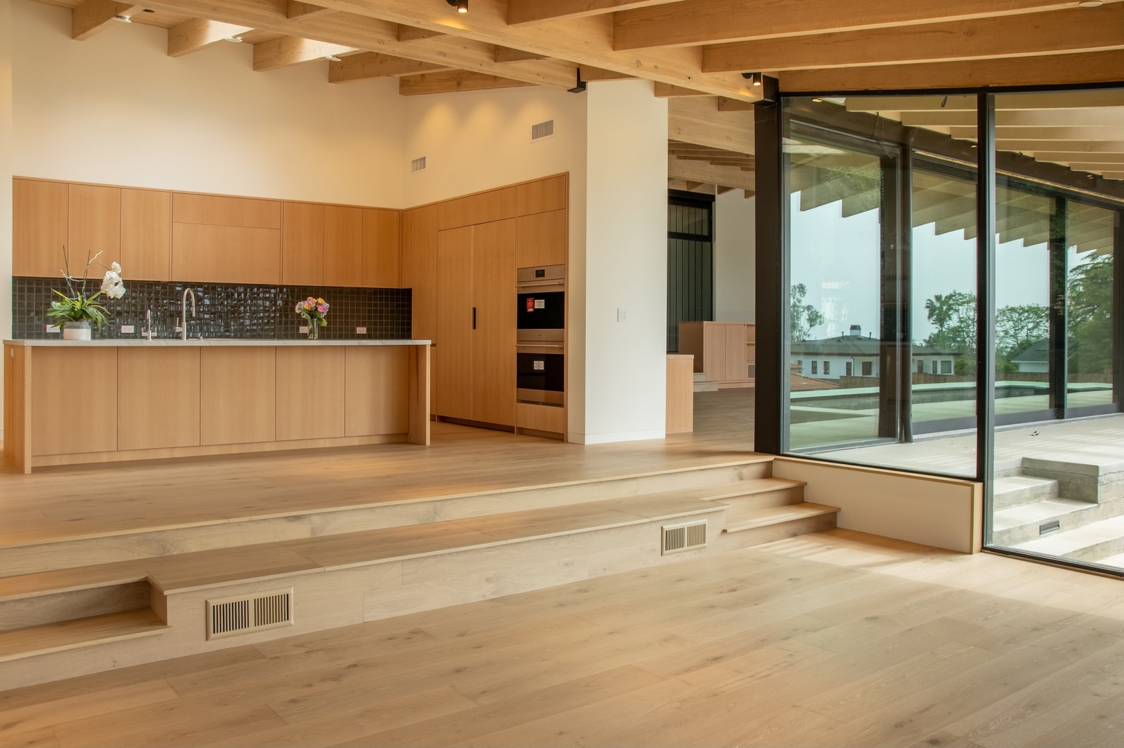 Open-plan living and kitchen with exposed wood beams and full-height glass walls — La Cañada